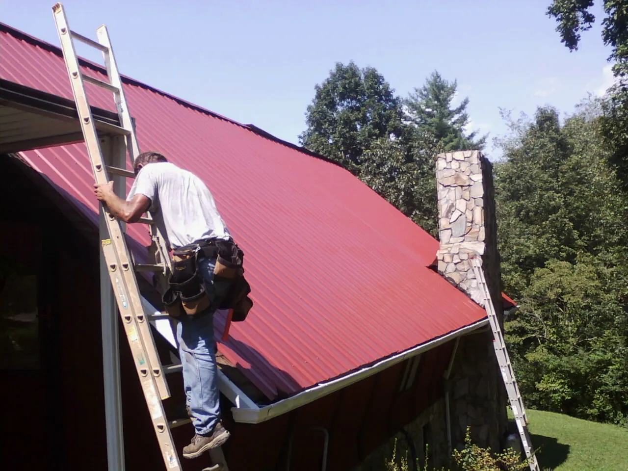 Roofer on a ladder beside a red metal roof and stone chimney — Soffit & Fascia Repair by Roofing Brothers in Kew Gardens